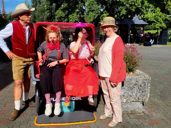 Vier Personen in bunten Kostümen posieren im Freien. Ein Mann im Cowboy-Outfit steht links neben einem kleinen roten Fahrzeug. Darin sitzen zwei Frauen, eine als Clown verkleidet. Rechts daneben steht eine Frau im Hut und lächelt.