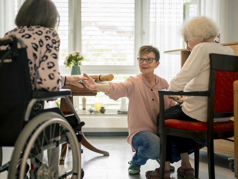 Eine Mitarbeiterin unterhält sich mit zwei Bewohnerinnen und reicht einer der beiden die Hand.