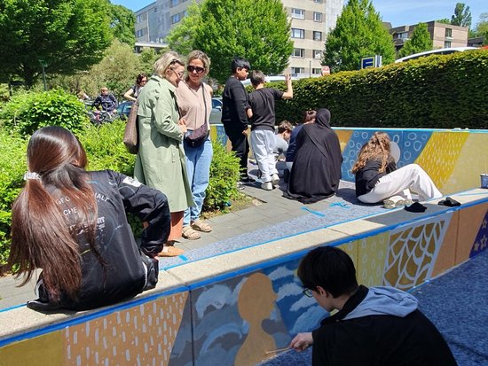 Eine Gruppe junger Menschen entspannt an einem sonnigen Tag auf einer farbenfroh bemalten Betonmauer im Freien. Im Vordergrund sitzen ein Mädchen mit langem Pferdeschwanz und ein Junge mit Brille. Im Hintergrund unterhalten sich weitere Personen vor grünen Hecken und Bäumen.