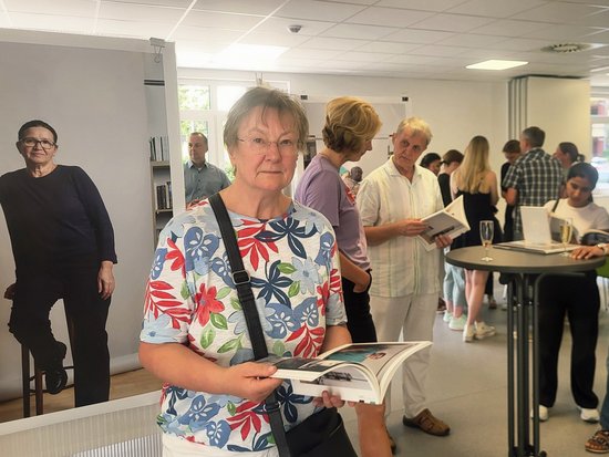 Eine Frau in einem blumigen Oberteil hält im Vordergrund ein offenes Buch. Im Hintergrund betrachten weitere Besucher an einem Tisch ebenfalls Bücher. Links steht eine Plakatwand mit einem lebensgroßen Foto einer Frau.