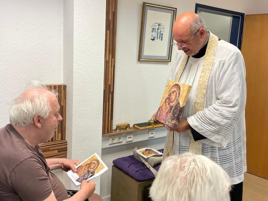 Ein Priester in liturgischen Gewändern steht und hält eine Ikone. Ein Mann im Rollstuhl und eine Frau halten ebenfalls religiöse Bilder in den Händen. Die Szene zeigt eine religiöse Zeremonie in einem Innenraum.