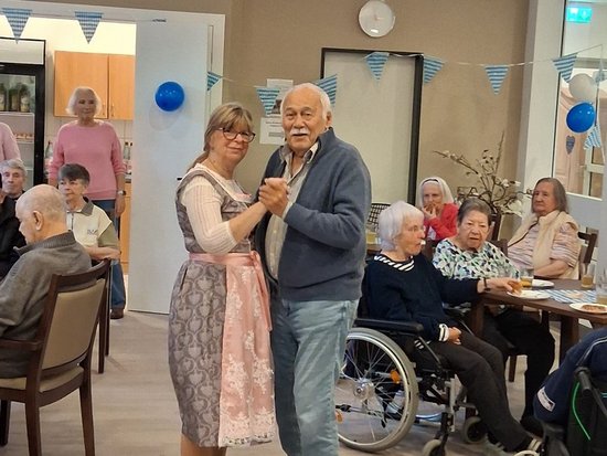 Ein älteres Paar tanzt Hand in Hand in einem hellen Gemeinschaftsraum. Im Hintergrund beobachten andere Senioren, teilweise im Rollstuhl, das Geschehen. Blaue und weiße Wimpelketten sowie Luftballons deuten auf eine festliche Veranstaltung in einer Pflegeeinrichtung hin.