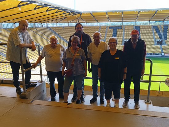 Sieben Personen posieren für ein Gruppenfoto auf einer Tribüne in einem Fußballstadion. Im Hintergrund sind gelbe Sitzreihen und der grüne Rasen zu erkennen.