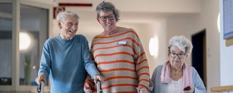 Haus St. Raphael Zwei Bewohnende spazieren mit einer Mitarbeiterin über den Flur. Sie halten sich an den Händen.