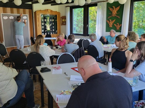 Ein Mann hält vor einer Tafel einen Vortrag für eine Gruppe von Erwachsenen, die an Tischen in einem hellen Raum mit Holzvertäfelung sitzen und zuhören.