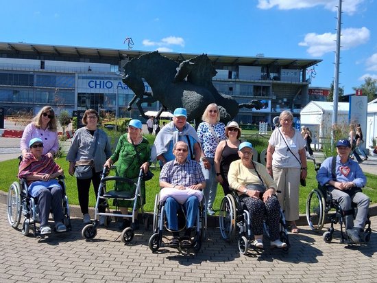 Eine Gruppe von Menschen, darunter mehrere Personen in Rollstühlen, posiert für ein Foto auf einem Platz. Im Hintergrund steht eine große Bronzestatue eines springenden Pferdes vor einem Gebäude mit der Aufschrift „CHIO" unter blauem Himmel.