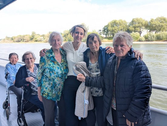 Sechs Frauen posieren auf einem Boot. Zwei sitzen im Rollstuhl, vier stehen. Eine jüngere Frau in der Mitte umarmt die älteren Damen. Im Hintergrund ist ein Fluss mit Bäumen zu sehen.