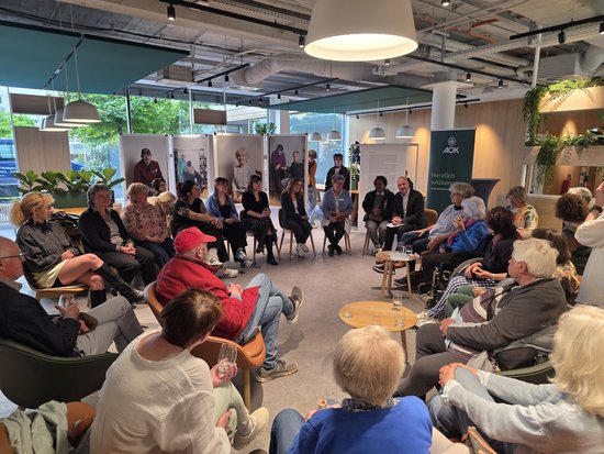 Eine Gruppe von Menschen sitzt in einem hellen, modernen Raum in einem großen Kreis. Im Hintergrund sind große Fenster, Pflanzen und Ausstellungstafeln zu sehen. Die Personen scheinen an einer Diskussion oder einem geselligen Treffen teilzunehmen.