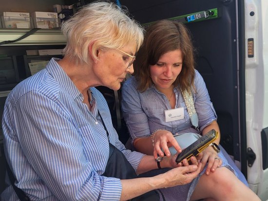 Zwei Frauen sitzen neben einem weißen Van mit dem Logo „Digitaler Engel". Eine jüngere Frau erklärt einer älteren Dame die Nutzung eines Tablets, auf das sie zeigt.